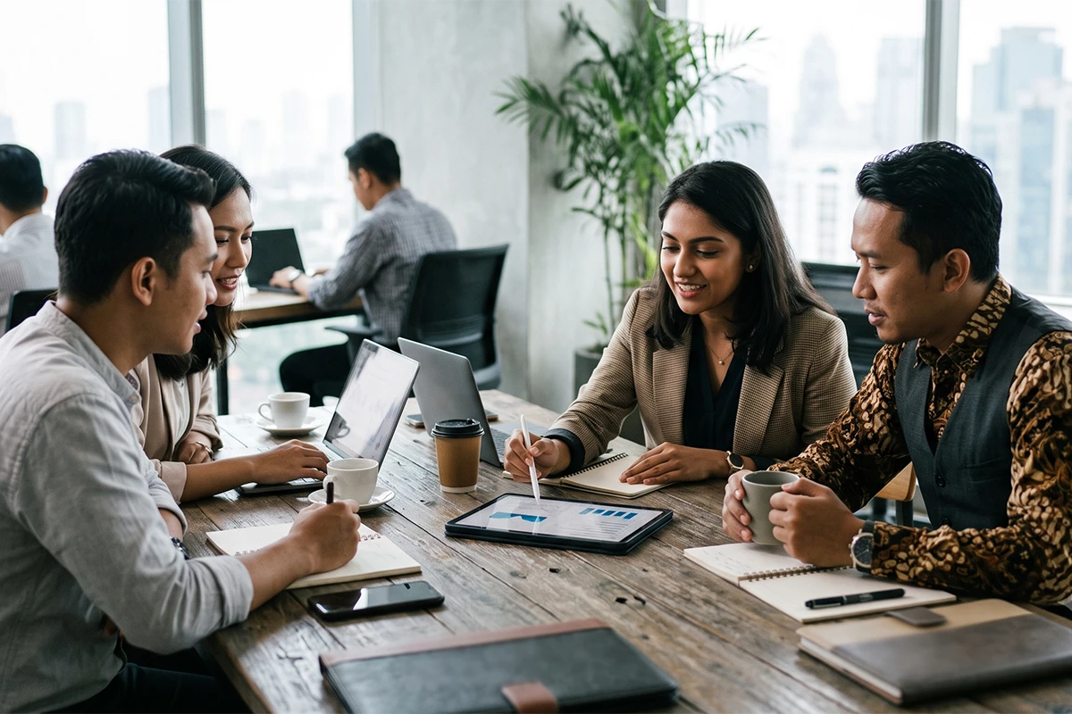 A group of four diverse professionals, including a man in a traditional batik shirt and a woman using a tablet stylus, discussing financial data around a rustic wooden table in a high-rise office.